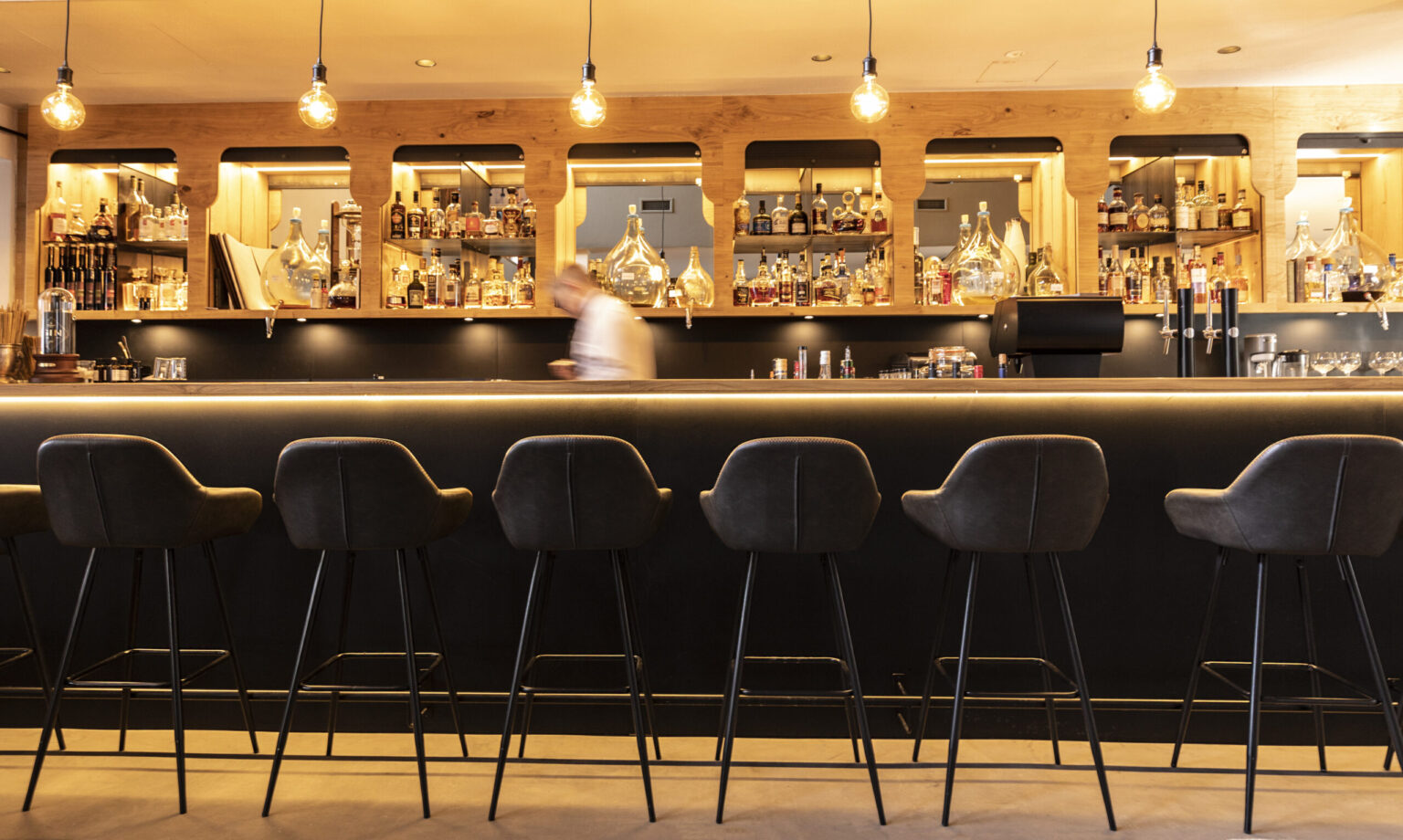 Forsthofgut 6.0 Moderne Gastronomiebarküche mit eleganter Bartheke und stilvollen Lederbarhockern. Die Regale aus Holz sind mit edlen Spirituosen gefüllt und die Beleuchtung sorgt für eine gemütliche Atmosphäre. FHE GASTRO bietet maßgeschneiderte Einrichtungen für anspruchsvolle Bars und Restaurants.