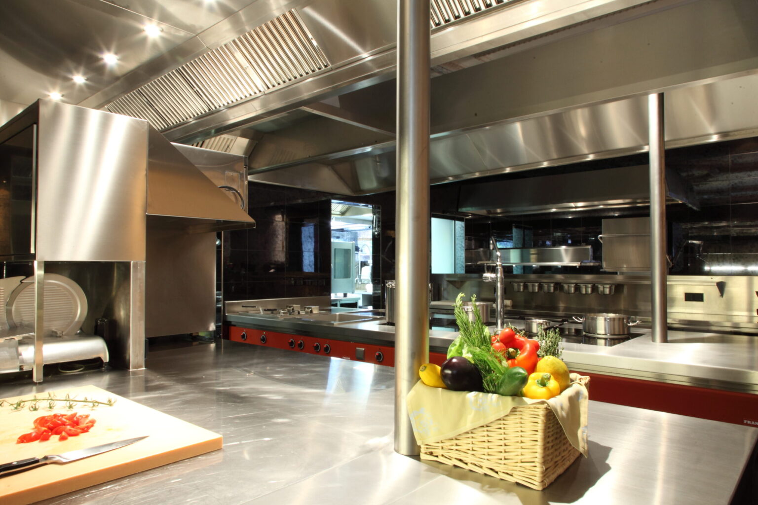 Hochwertige Edelstahlküche mit einer offenen Kochstation, die verschiedene Töpfe und einen Korb mit frischen Gemüsezutaten wie Paprika, Tomaten und Auberginen zeigt, daneben eine Induktionskochstelle und moderne Dunstabzugshauben