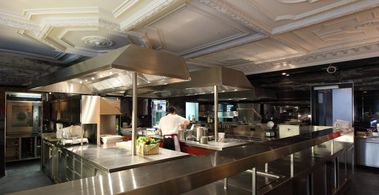 Ein professioneller Koch bereitet eine Mahlzeit in einer hochmodernen Edelstahlküche zu, umgeben von professionellen Küchengeräten wie Induktionskochfeldern, Dunstabzugshauben und verschiedenen Töpfen und Pfannen