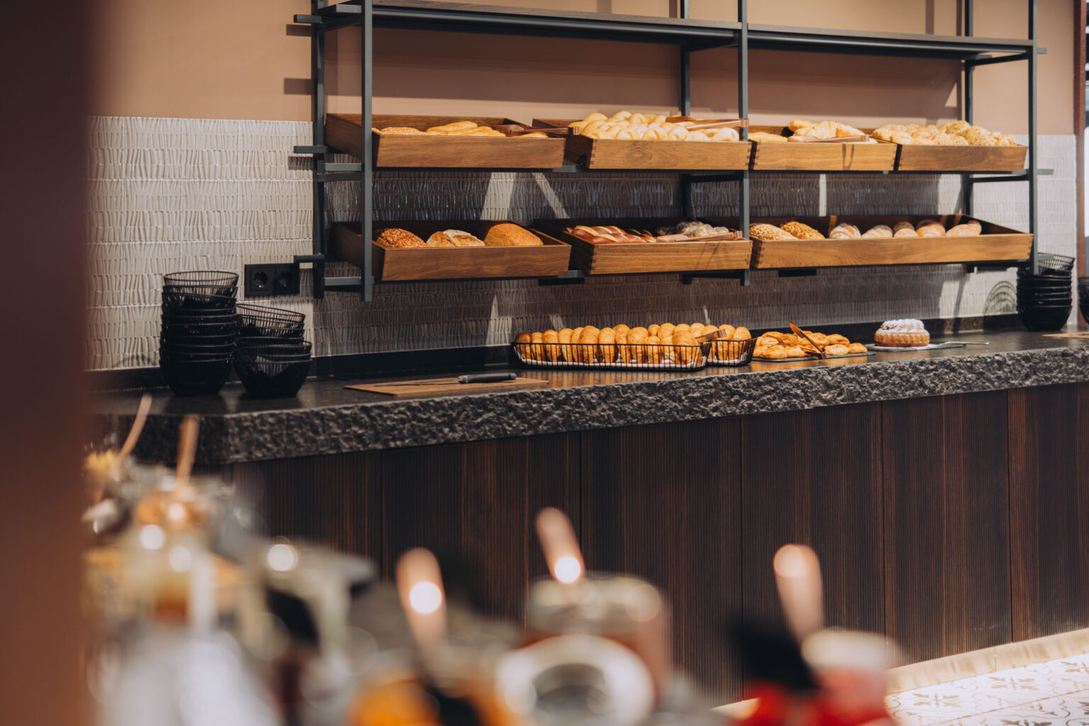 Fernblick_Bartholomäberg Buffet 2024 (4) Brotbuffet von FHE GASTRO mit einer großen Auswahl an frischem Gebäck und Brotsorten, präsentiert in stilvollen Holzregalen. Eine rustikale Steinoberfläche verleiht dem Bereich ein warmes Ambiente.