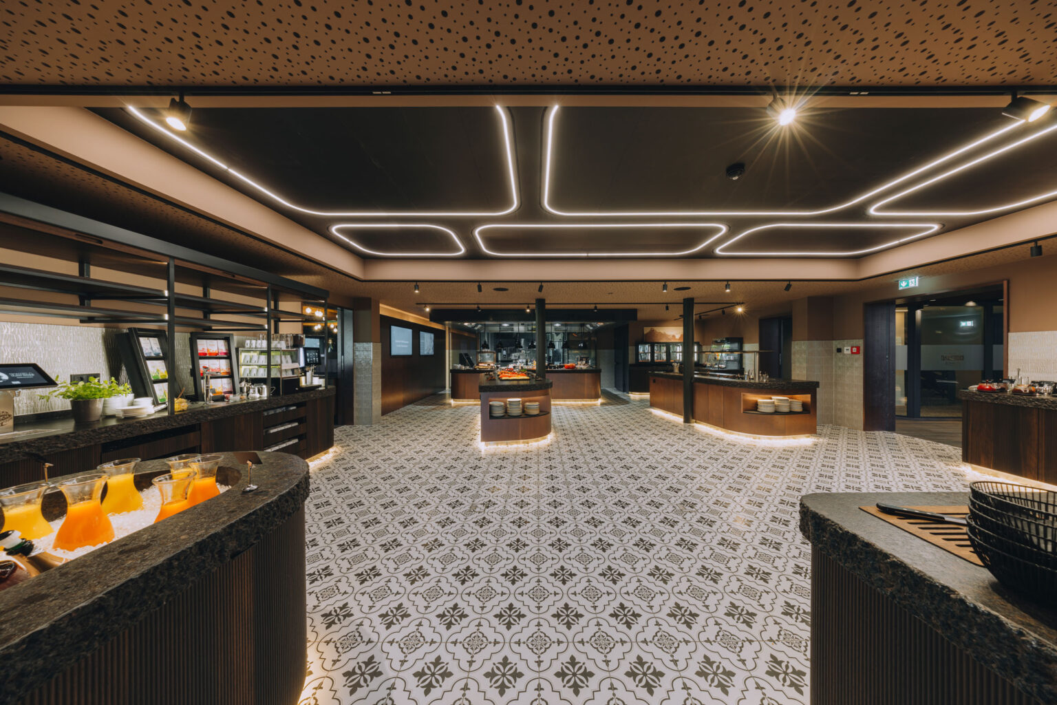 Fernblick_Bartholomäberg Buffet 2024 (8) Weitläufiger Buffetbereich von FHE GASTRO mit elegantem Lichtdesign in der Decke und einem kunstvollen Fliesenbodenmuster. Verschiedene Stationen für Getränke, Salate und warme Speisen umrahmen den Raum.