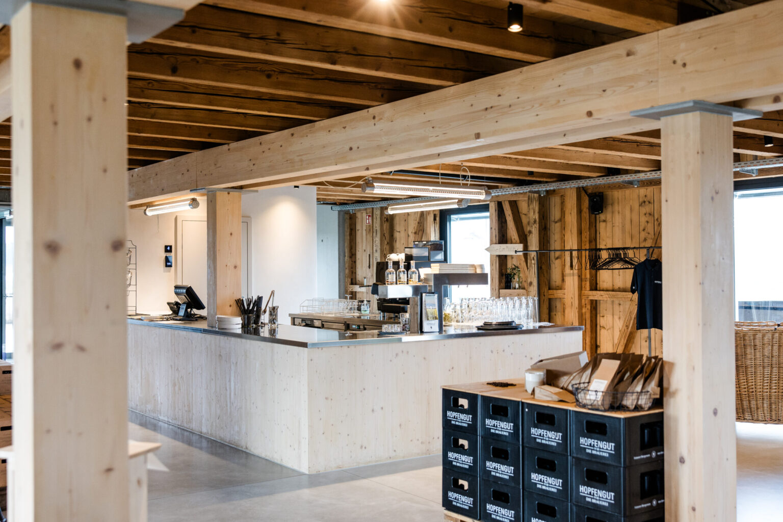 Hopfengut_2024 (3) Modernes, rustikales Café von FHE GASTRO im Hopfengut-Stil mit Holzbalken und offenen Strukturen. Der Tresen ist schlicht gestaltet, während Holzstapeln und Kisten dem Raum eine ländliche Note verleihen.