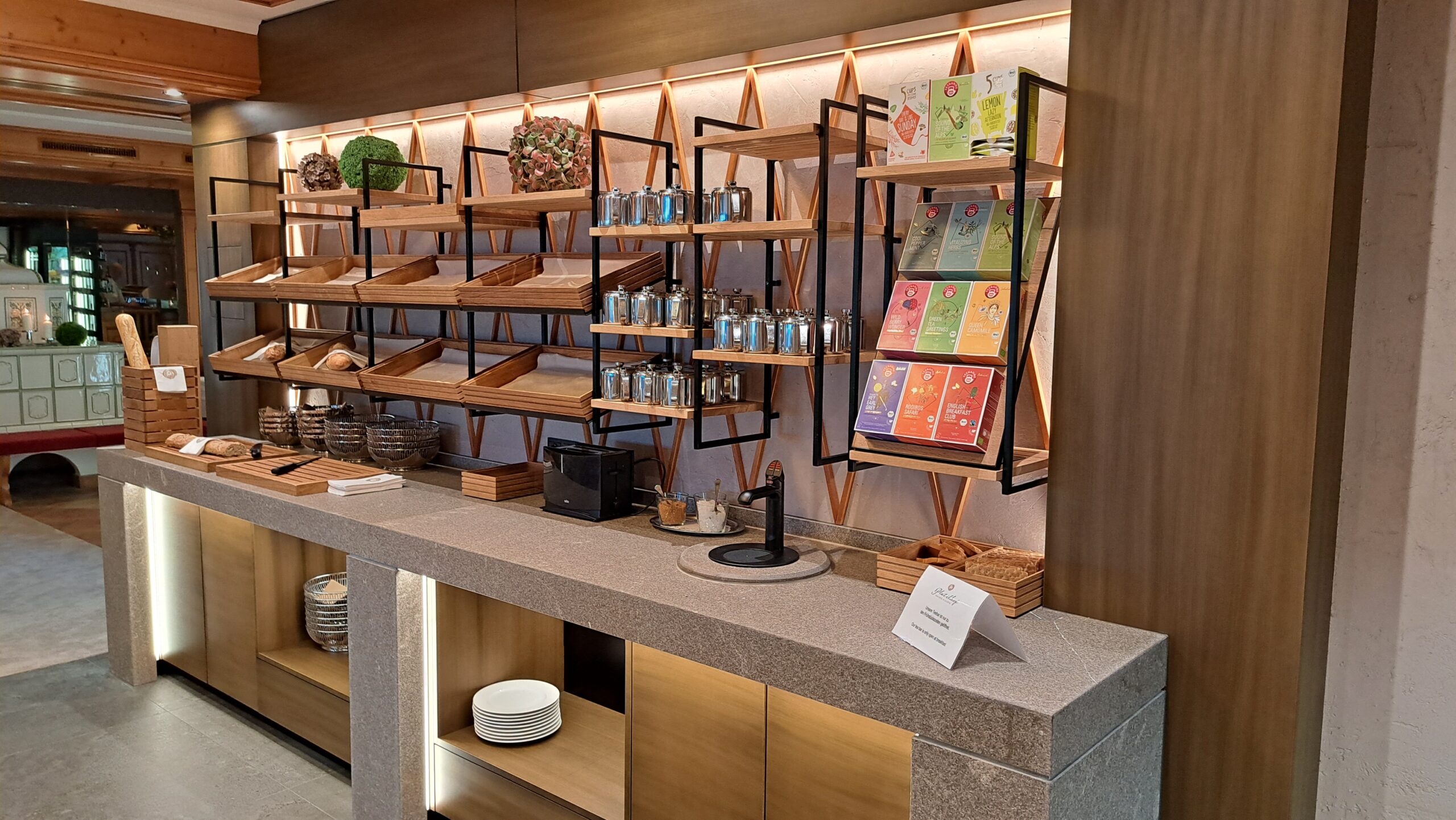 Modernes Buffet von FHE GASTRO in einem eleganten Gastronomiebereich. Die Theke kombiniert funktionales Design mit hochwertigem Granit und warmen Holzelementen. Auf den Regalen sind stilvolle Vorratsbehälter und Teekartons aufgereiht. FHE GASTRO liefert maßgeschneiderte Möbel und Einrichtungen, die sowohl ästhetisch als auch praktisch für gastronomische Betriebe sind.