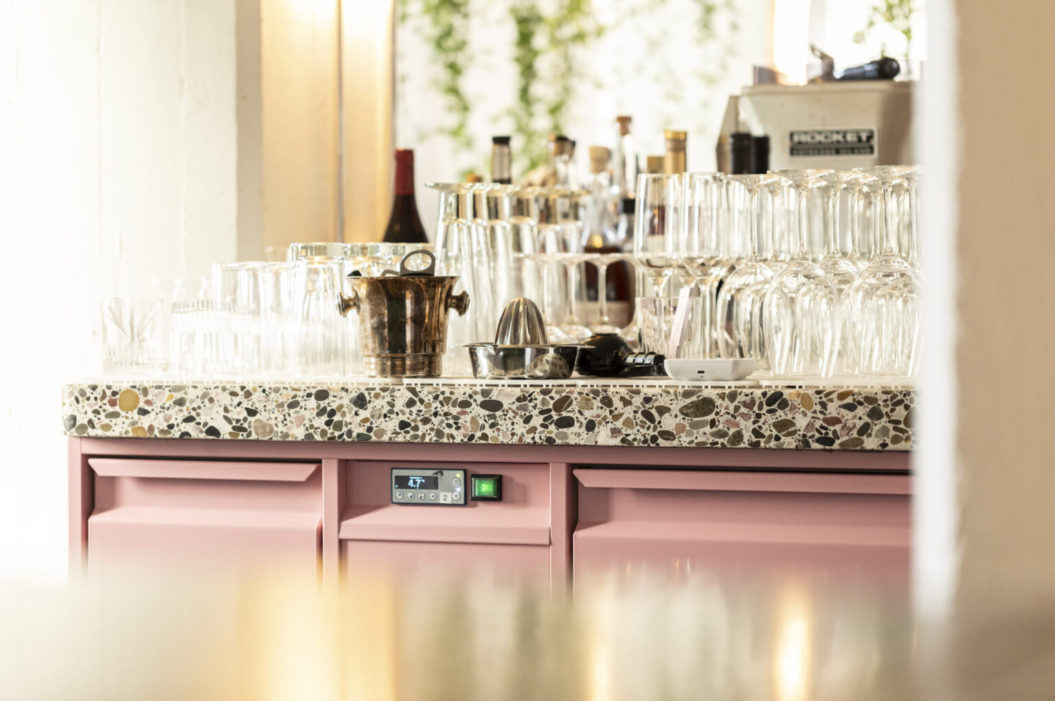 Detailansicht einer Bar von FHE GASTRO mit Gläsern, Barutensilien und einer Terrazzo-Arbeitsplatte. Das innovative Design der Küchenschränke in Pink und das professionelle Barzubehör unterstreichen das moderne Ambiente für Gastronomiebetriebe. FHE GASTRO bietet individuelle Möbel und Lösungen, die Funktionalität und Stil vereinen.