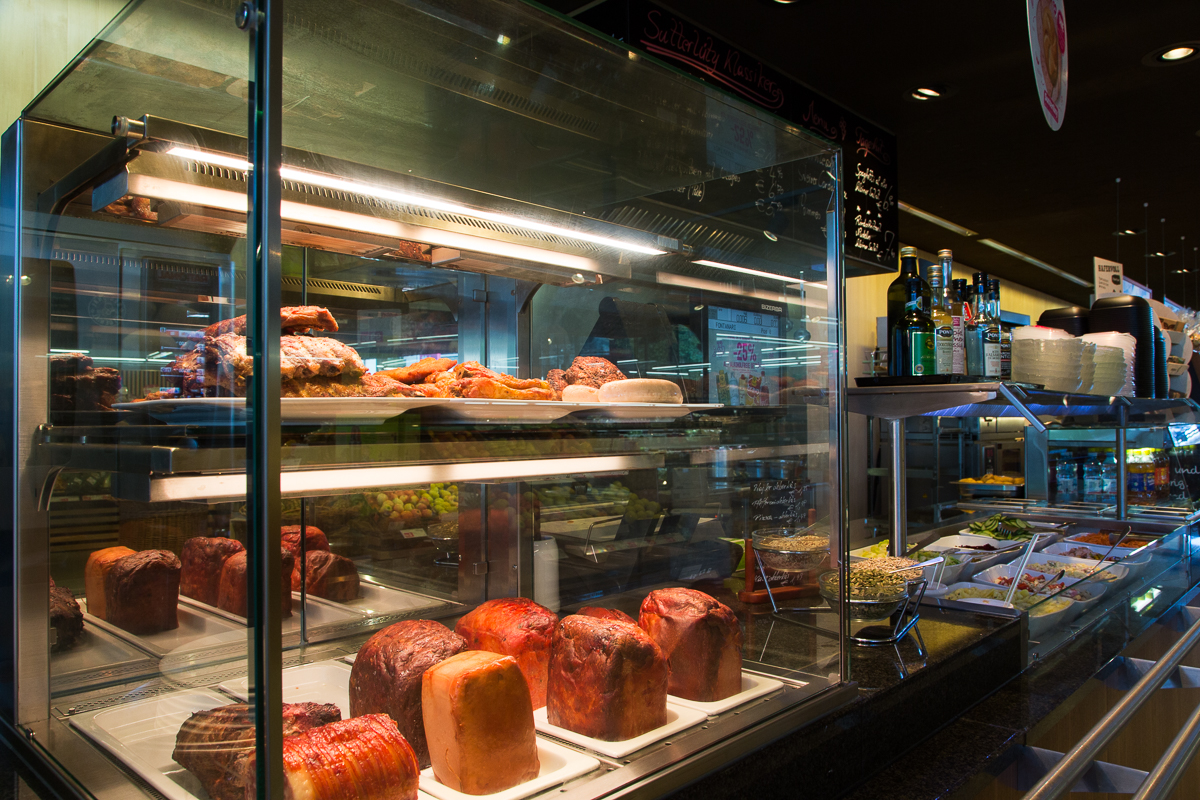 Theke im Sutterlüty Supermarkt mit einer Auswahl an frisch zubereiteten Fleisch- und Gemüsegerichten in Vitrinen. FHE GASTRO bietet hochwertige Gastronomieeinrichtungen für die Präsentation und den Verkauf von frischen Lebensmitteln.
