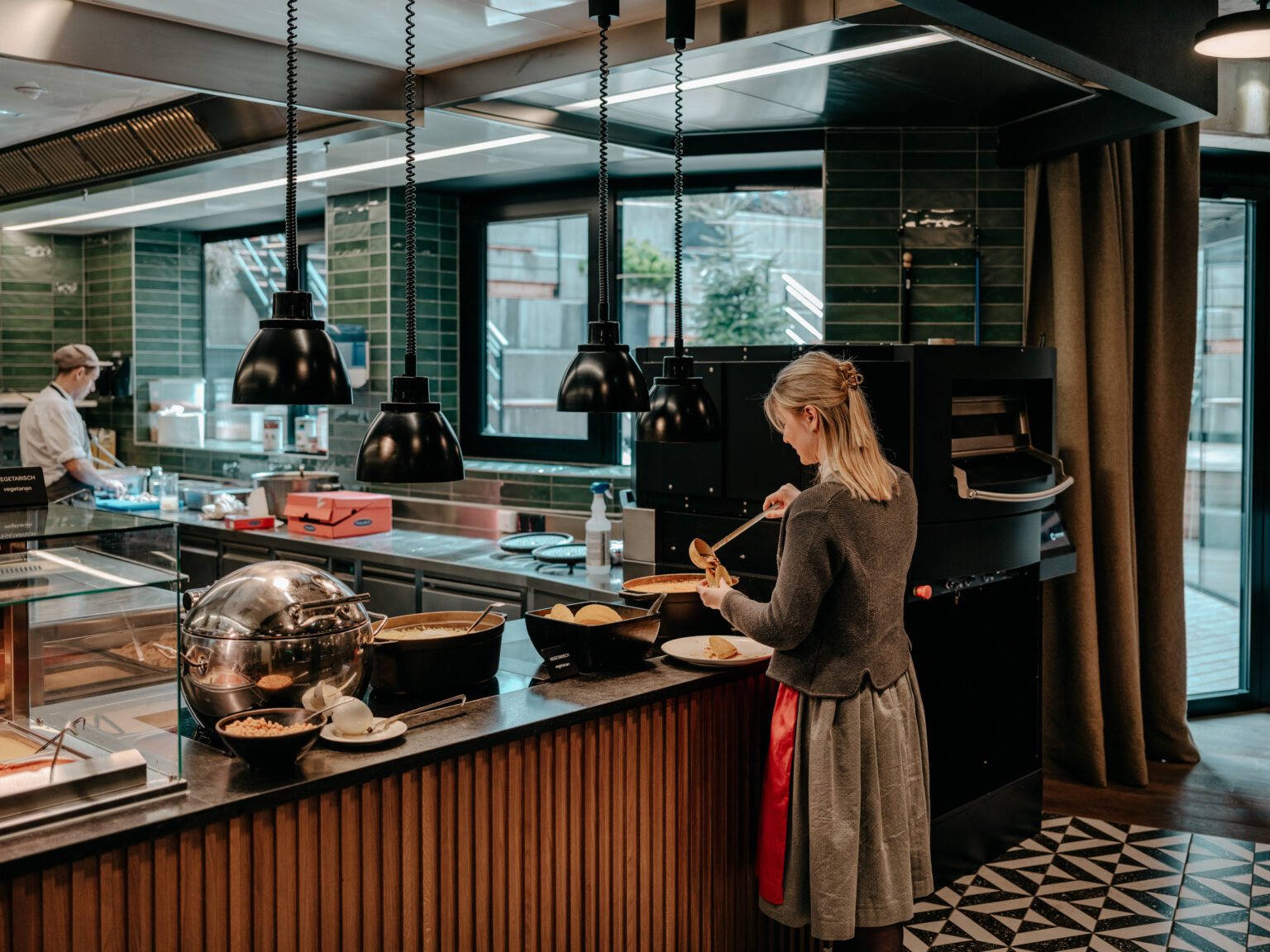 240312_Forsthofgut_patrese-131 Moderne, offene Küchenstation mit hochwertiger Ausstattung für die Gastronomie. Ein Koch bereitet in der professionellen Küche frische Gerichte zu, umgeben von Edelstahlflächen und praktischen Gastro-Geräten. FHE GASTRO bietet maßgeschneiderte Küchenlösungen für Restaurants und Hotels, die sowohl Funktionalität als auch ästhetisches Design kombinieren