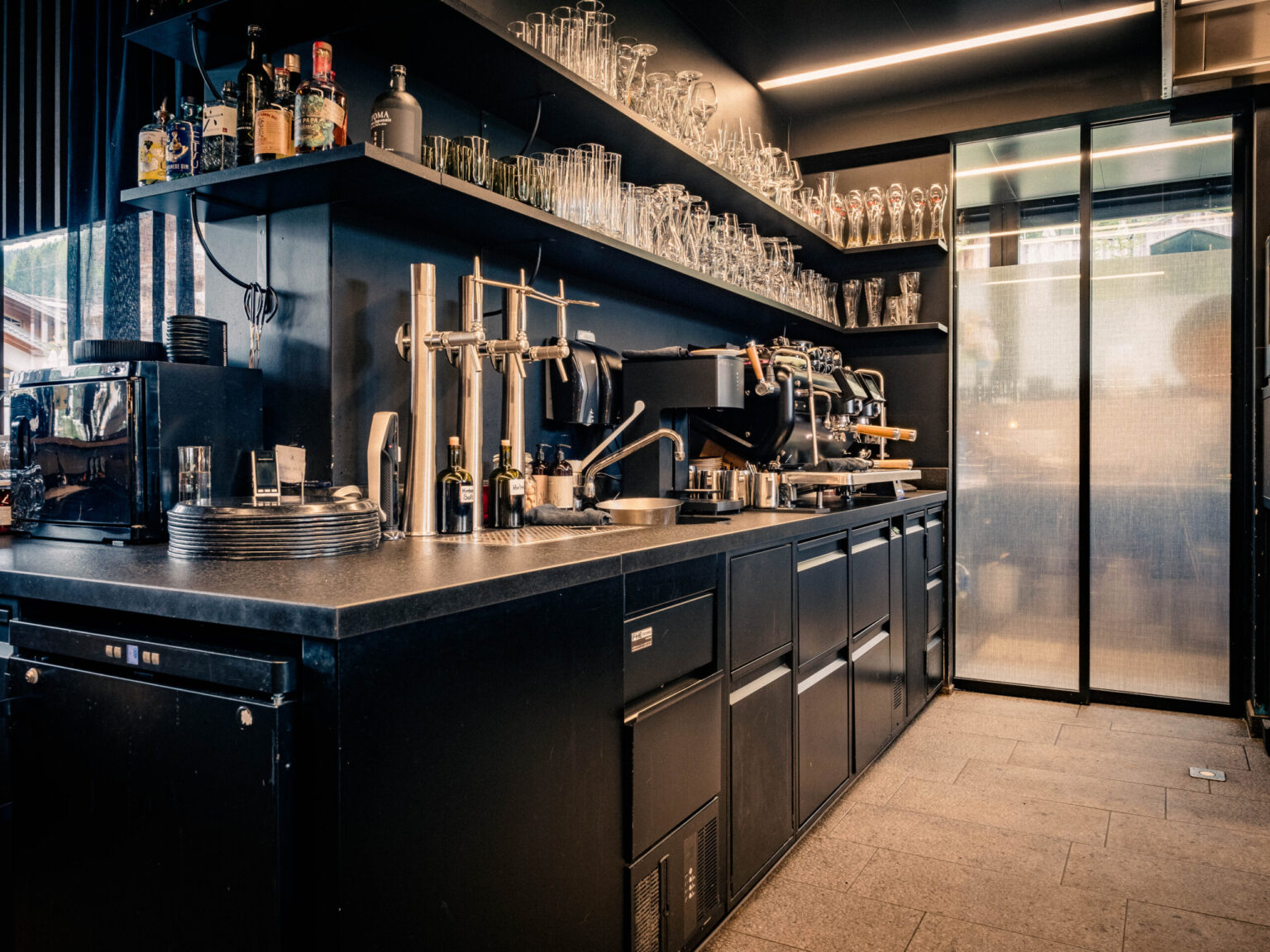 MIZUMI_Forsthofgut-April_2024-422-HDR Voll ausgestatteter Barbereich mit professionellen Geräten von FHE GASTRO, einschließlich einer modernen Kaffeemaschine und Spirituosenregalen.